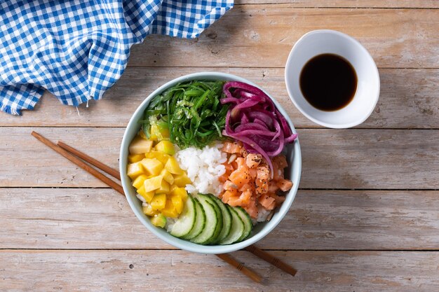 Poke bowl con arroz, salmón, pepino, mango, cebolla, ensalada de wakame, semillas de amapola y semillas de girasol en la mesa de madera.