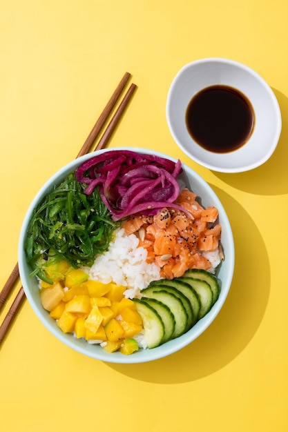 Poke bowl con arroz, salmón, pepino, mango, cebolla, ensalada de wakame, semillas de amapola y semillas de girasol sobre fondo amarillo.