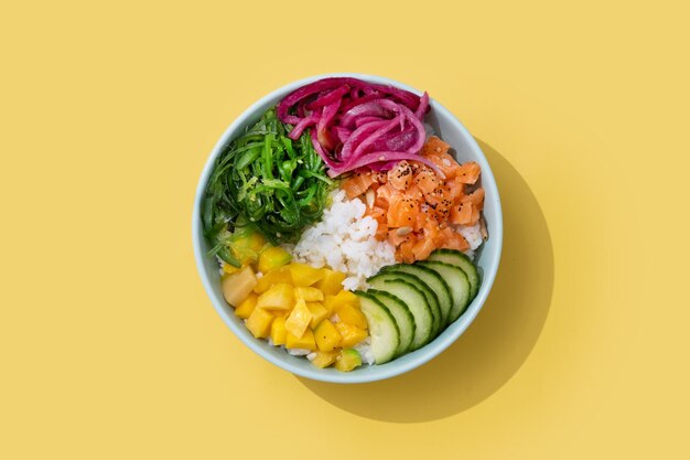 Poke bowl con arroz salmónpepinomangocebollawakame ensalada semillas de amapola y semillas de girasol aisladas sobre fondo amarilloxA