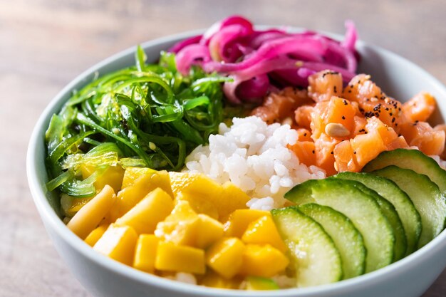 Poke bowl con arroz salmónpepinomangocebollawakame ensalada semillas de amapola y semillas de girasol sobre fondo de madera