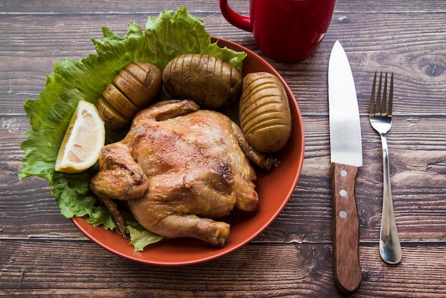 Pollo asado entero en un bol con papa; cuchillo y tenedor en mesa de madera