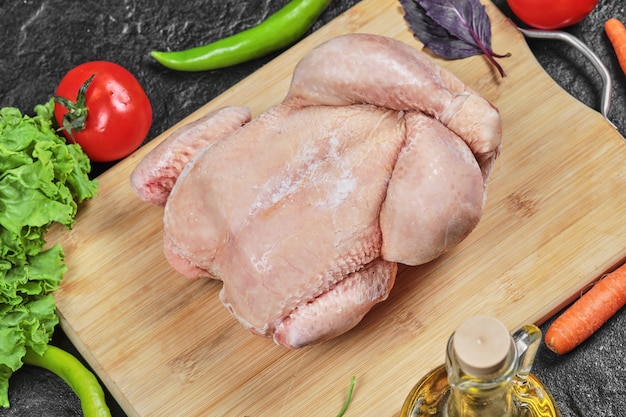 Pollo entero crudo sobre tabla de madera con lechuga, pimientos, aceite y tomates