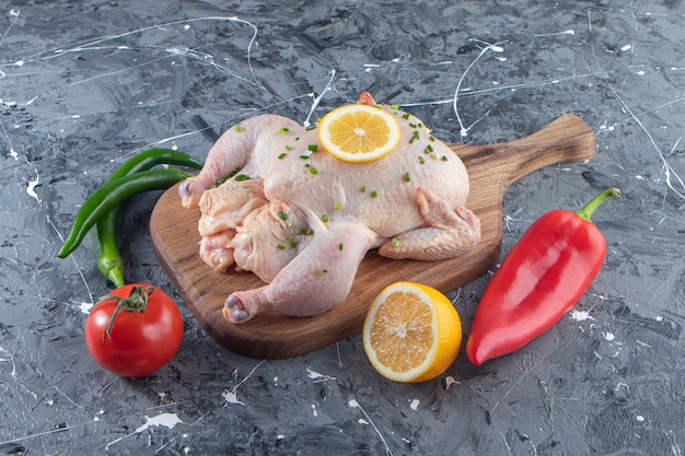 Pollo entero marinado crudo sobre una tabla de cortar junto a verduras, sobre la superficie de mármol.