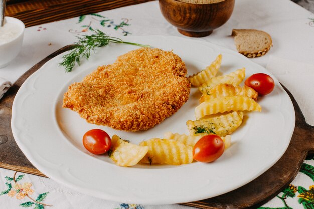 pollo frito junto con papas tomate rojo dentro de un plato blanco sobre escritorio marrón