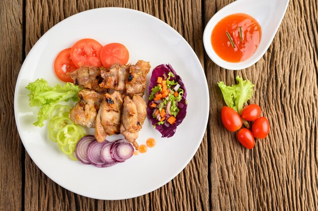 Pollo a la parrilla en un plato blanco con una ensalada, tomates, chiles cortados en trozos y salsa en la mesa de madera.