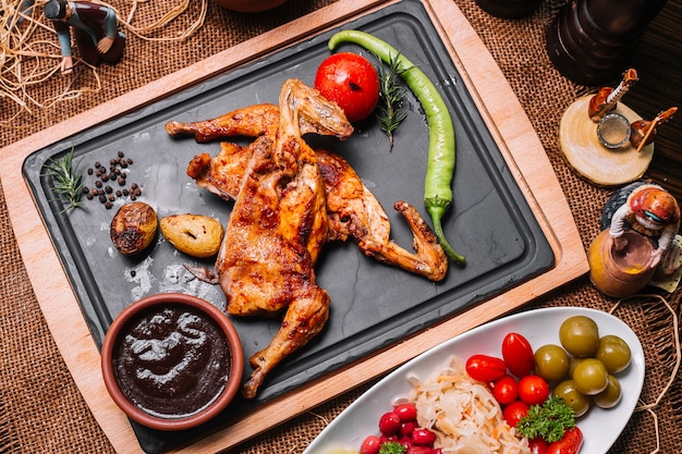 Pollo a la parrilla sobre tabla de madera con salsas de tomate, pimiento, papa, encurtidos, vista superior