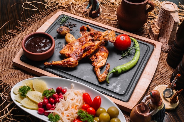 Pollo a la parrilla sobre tabla de madera con tomate papas salsas de pimienta encurtidos vista lateral