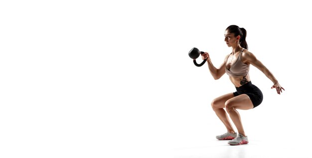 Se pone en cuclillas con peso. Entrenamiento de la atleta femenina profesional caucásica aislada en la pared blanca. Mujer musculosa y deportiva. Concepto de acción, movimiento, juventud, estilo de vida saludable. Copyspace para anuncio.