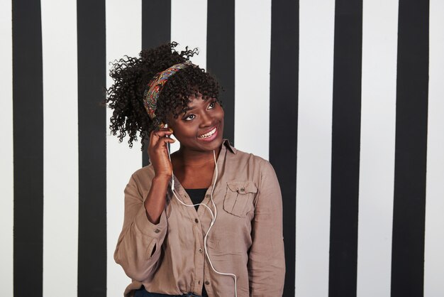 Poniendo auriculares en el oído y me sorprendió la música. Niña afroamericana sonrió se encuentra en el estudio con líneas verticales blancas y negras en el fondo