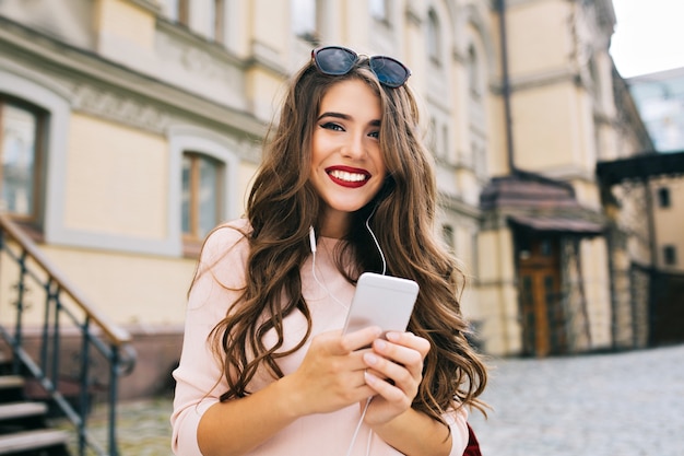 Portraut de linda chica con pelo largo y rizado y teléfono en manos sonriendo en la ciudad en el fondo del edificio