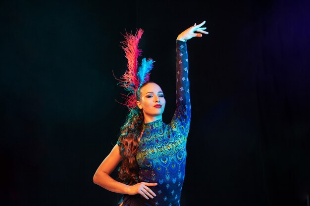 Posando. Hermosa mujer joven en carnaval, elegante disfraz de mascarada con plumas en pared negra en luz de neón. Copyspace para anuncio. Celebración de fiestas, baile, moda. Tiempo festivo, fiesta.