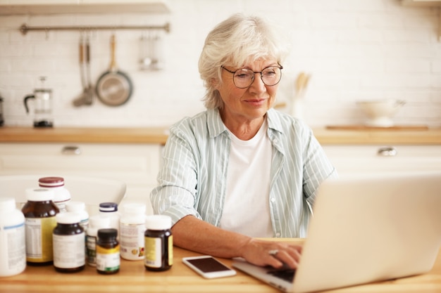 Positiva hermosa jubilada de pelo gris con gafas que elige un estilo de vida saludable, sentada en la cocina con suplementos dietéticos, escribiendo en el teclado en la computadora portátil, escribiendo revisión a través de la tienda en línea