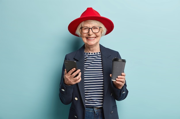 Positiva hermosa mujer pelirroja con gafas, usa teléfono celular, envía mensajes a través de una aplicación multimedia, navega por las redes sociales, toma un descanso para tomar café, sostiene una taza de bebida desechable, aislada en azul