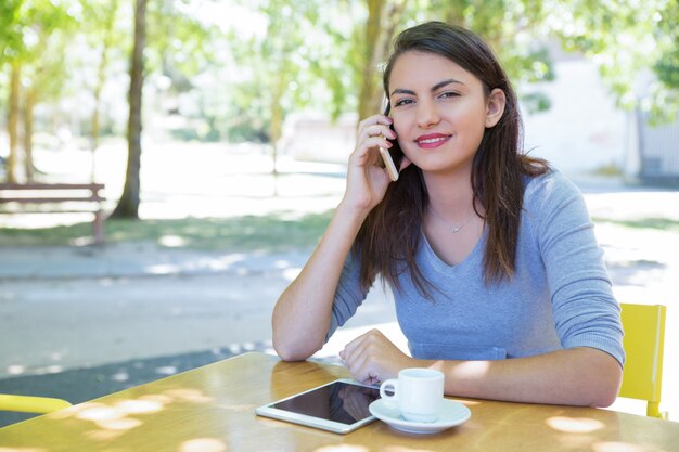 Positiva jovencita hablando por teléfono en la mesa de café en el parque