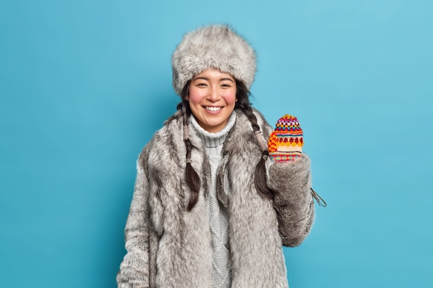 Positiva morena joven asiática lleva abrigo de piel y sombrero olas mano en guantes de punto sonríe alegremente disfruta de vacaciones de invierno o vacaciones aisladas sobre pared azul