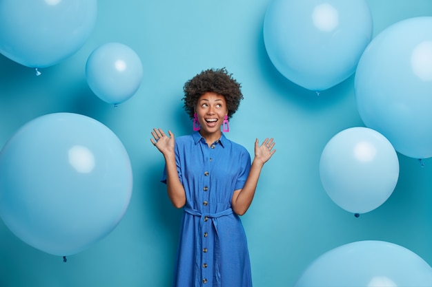 Positiva mujer afroamericana despreocupada lista para la celebración, vestida con ropa festiva, posa contra globos azules