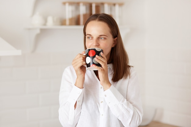 Positiva mujer de pelo oscuro con camiseta blanca de estilo casual, tomando café o té por la mañana, posando con un juego de cocina de fondo, mirando a otro lado con expresión soñadora.