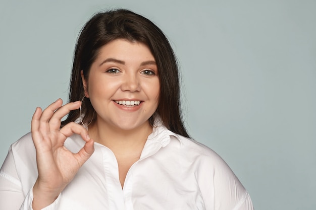 Positividad, éxito, aprobación y lenguaje corporal. Alegre mujer joven exitosa con mejillas regordetas sonriendo con confianza, haciendo un gesto bien mientras posa