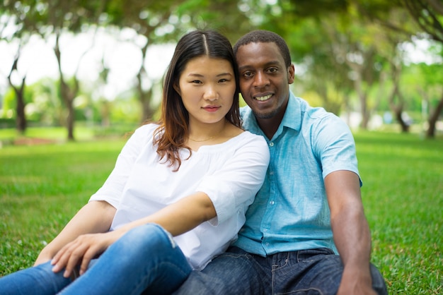 Positivo calma hermosa joven pareja interracial mirando a cámara mientras descansa sobre hierba