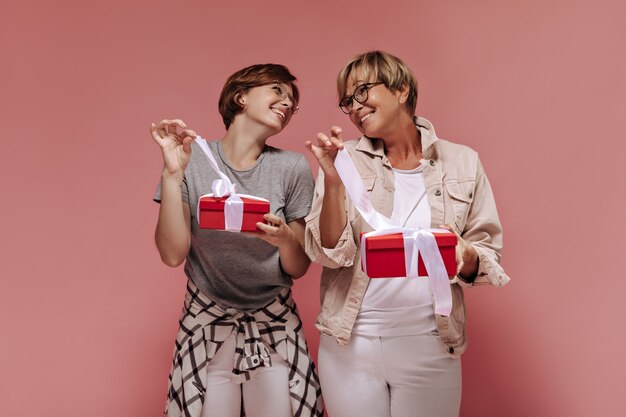 Positivo dos mujeres con peinado moderno corto y anteojos de moda en traje ligero mirando y desatando cintas en cajas de regalo sobre fondo rosa.