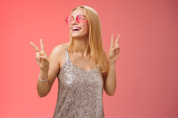 Positivo encantador europeo despreocupado elegante novia divirtiéndose en la pista de baile muestran signos de paz de victoria mirar hacia arriba sonrisa feliz en gafas de sol glamour vestido brillante disfrutar de celebración, fondo rojo.