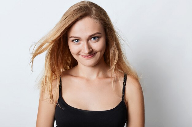 Positivo joven hermosa mujer con exuberante cabello rubio posa contra estudio blanco