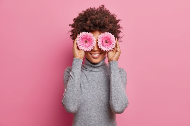 Positivo joven de piel oscura disfruta del día de primavera, tiene flores de gerbera rosa en los ojos