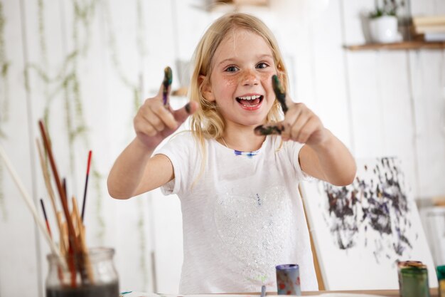 Positivo y lleno de alegría, sonriendo con dientes rubia niña europea señalando con los dedos en la pintura.