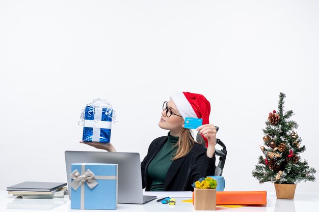 Positivo, mujer de negocios, con, santa claus, sombrero, y, llevando lentes, sentado, en una mesa, tenencia, navidad, regalo, y, tarjeta bancaria, en la oficina colección de imágen