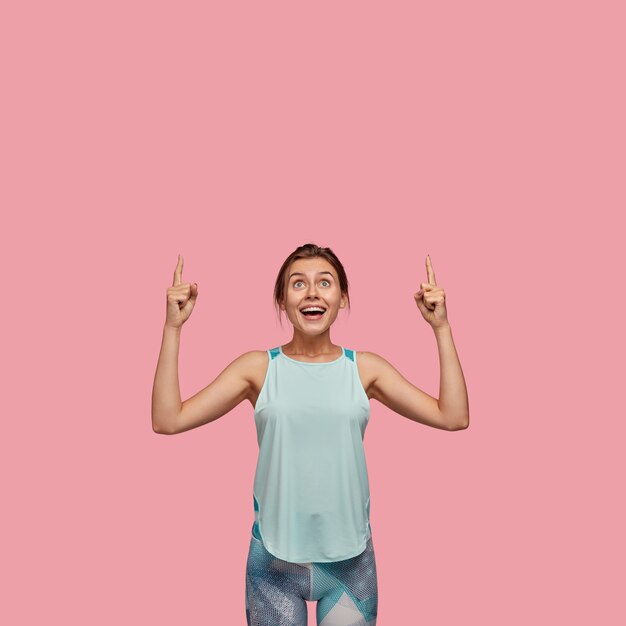 Positve mujer europea con expresión alegre, apunta con ambos dedos índices hacia arriba, vestida con chaleco informal y leggings, modelos sobre pared rosa. Concepto de publicidad. ¡Mira el techo!