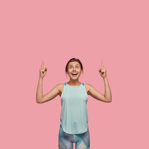 Positve mujer europea con expresión alegre, apunta con ambos dedos índices hacia arriba, vestida con chaleco informal y leggings, modelos sobre pared rosa. Concepto de publicidad. ¡Mira el techo!