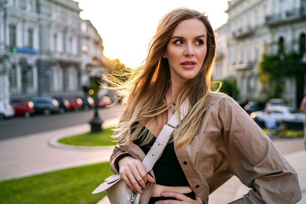 Postrait informal de la ciudad al aire libre de una elegante mujer rubia caminando sola divirtiéndose al atardecer