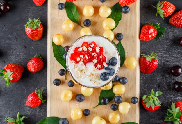 Postre con fresa, arándano, cereza, hojas en un jarrón sobre tabla gris y tabla de cortar, plano.