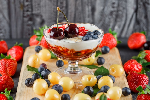 Postre con fresa, arándano, cereza, tabla de cortar en un jarrón sobre superficie gris y madera, vista de ángulo alto.