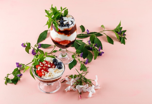 Postre con fresas, arándanos, nueces, menta, ramas de flores en copa y florero en superficie rosa, vista de ángulo alto.