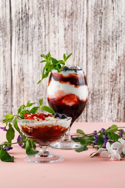 Postre con fresas, arándanos, nueces, menta, ramas de flores en copa y jarrón sobre superficie rosada y sucia, vista lateral.