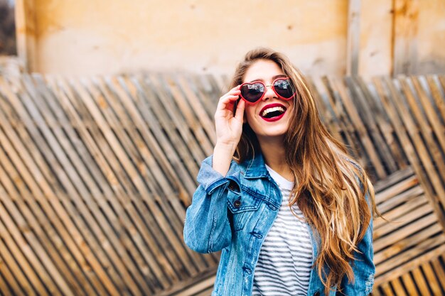 Postrtait de primer plano de modelo femenino magnífico en chaqueta de mezclilla retro, sosteniendo gafas de sol y mirando hacia arriba. Mujer joven sensual con el pelo largo hermoso que presenta con mucho gusto delante de la valla de madera inusual.