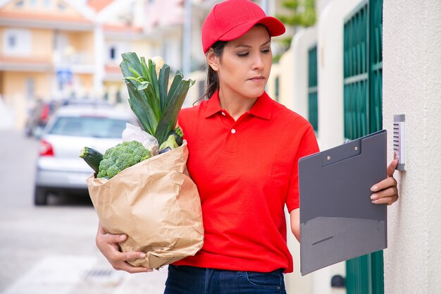 Postwoman bastante joven que sostiene la bolsa de papel y el timbre de la puerta. Repartidora morena confiada en uniforme rojo haciendo su trabajo y entregando el pedido a pie. Servicio de entrega de alimentos y concepto de correo.