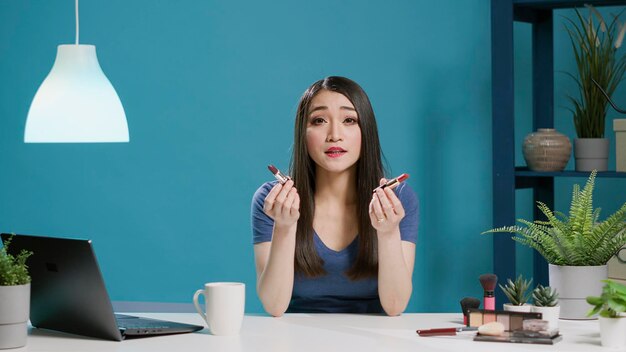 POV de influenciador asiático haciendo recomendación de pintalabios en el estudio, filmando revisión de maquillaje con productos cosméticos en cámara. Blogger femenina grabando video para redes sociales. Disparo de trípode.