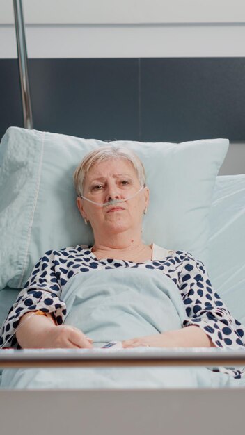 POV de mujer anciana con enfermedad hablando por videollamada para comunicación remota, tendido en la cama de la sala de hospital. Paciente mayor con tubo de oxígeno mediante conferencia en línea y mirando a la cámara