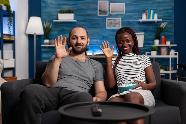 POV de una pareja joven interracial saludando en una videoconferencia mientras sostiene un tazón de palomitas de maíz. Personas que hablan con familiares a través de la tecnología de comunicación de videollamadas mientras están sentadas en la sala de estar.