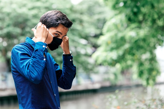 Precaución contra la contaminación. Hombre joven con cabello oscuro, con camisa azul mirando a otro lado, mientras usa máscara médica, de pie al aire libre. Seguridad, coronavirus, concepto pandémico