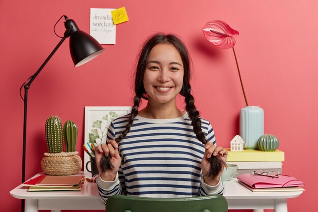 Preciosa colegiala se divierte mientras se prepara para los exámenes, tiene dos largas trenzas, sonríe feliz, vestida con un suéter a rayas, planea el horario para el próximo mes