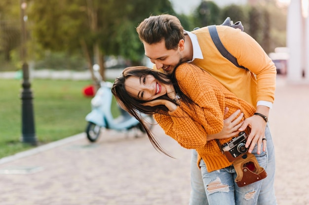 Preciosa modelo femenina en jeans rotos jugando con su esposo después de la sesión de fotos en el parque de otoño. Chica dichosa en suéter de punto con cámara divirtiéndose con novio disfrutando el fin de semana juntos