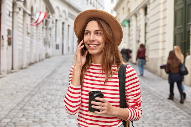 Precioso turista sonriente pasea por la ciudad, tiene una conversación telefónica, tiene café para llevar, se concentra en algún lugar en la distancia