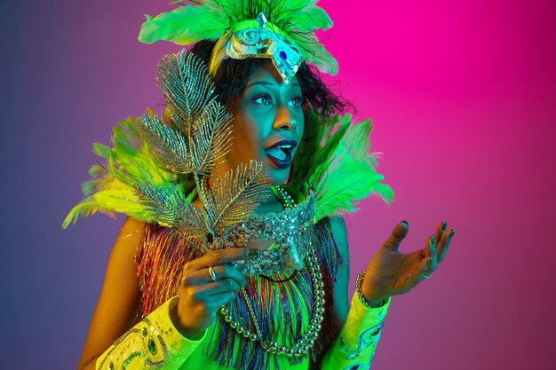 Preguntado. Hermosa mujer joven en carnaval, elegante disfraz de mascarada con plumas bailando en la pared degradada en neón. Concepto de celebración navideña, tiempo festivo, baile, fiesta, diversión.