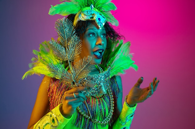Preguntado. Hermosa mujer joven en carnaval, elegante disfraz de mascarada con plumas bailando en la pared degradada en neón. Concepto de celebración navideña, tiempo festivo, baile, fiesta, diversión.