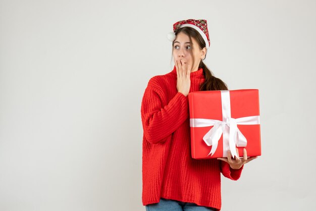 Se preguntó niña con gorro de Papá Noel poniendo la mano en la boca sosteniendo un regalo en blanco