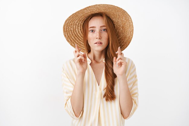 Preocupada, concentrada y guapa chica pelirroja con pecas en un lindo sombrero de paja de verano y una blusa a rayas cruzando los dedos para que tenga buena suerte, luciendo preocupada y preocupada rezando, pidiendo deseos