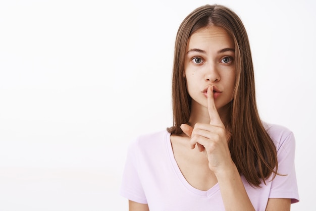 Preocupada encantadora y linda jovencita con cabello castaño corto mirando preocupada y gentil diciendo shh mostrando gesto de silencio con el dedo índice sobre la boca